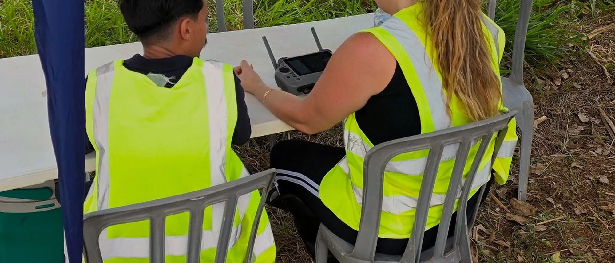 Dois operadores de colete refletivo, de costas, sentados à mesa de apoio sob a tenda azul na beira da lavoura, com o controle do DJI Agras sobre a mesa