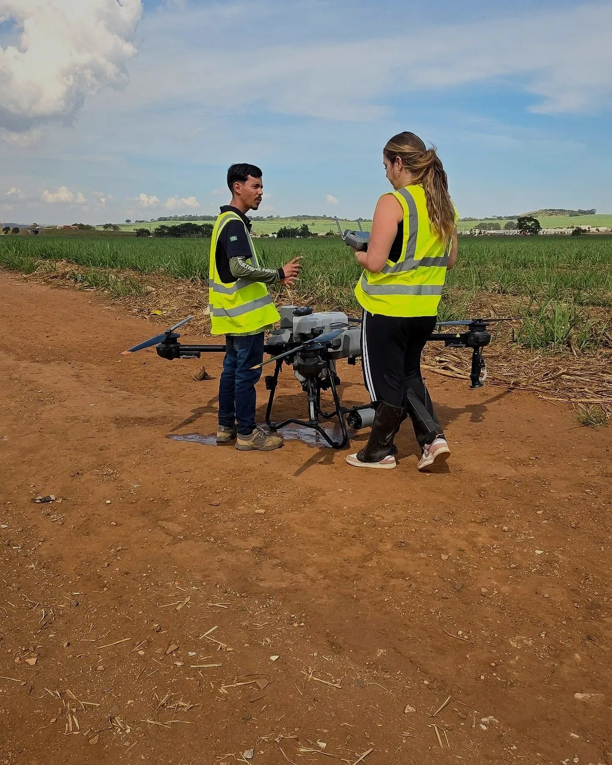 Dois alunos de colete conversam ao lado de um drone DJI Agras pousado no campo, antes do voo