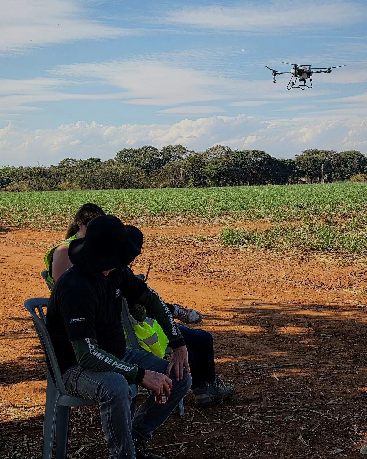 Instrutor e aluno de costas, sentados na sombra, acompanham o DJI Agras em voo sobre a lavoura