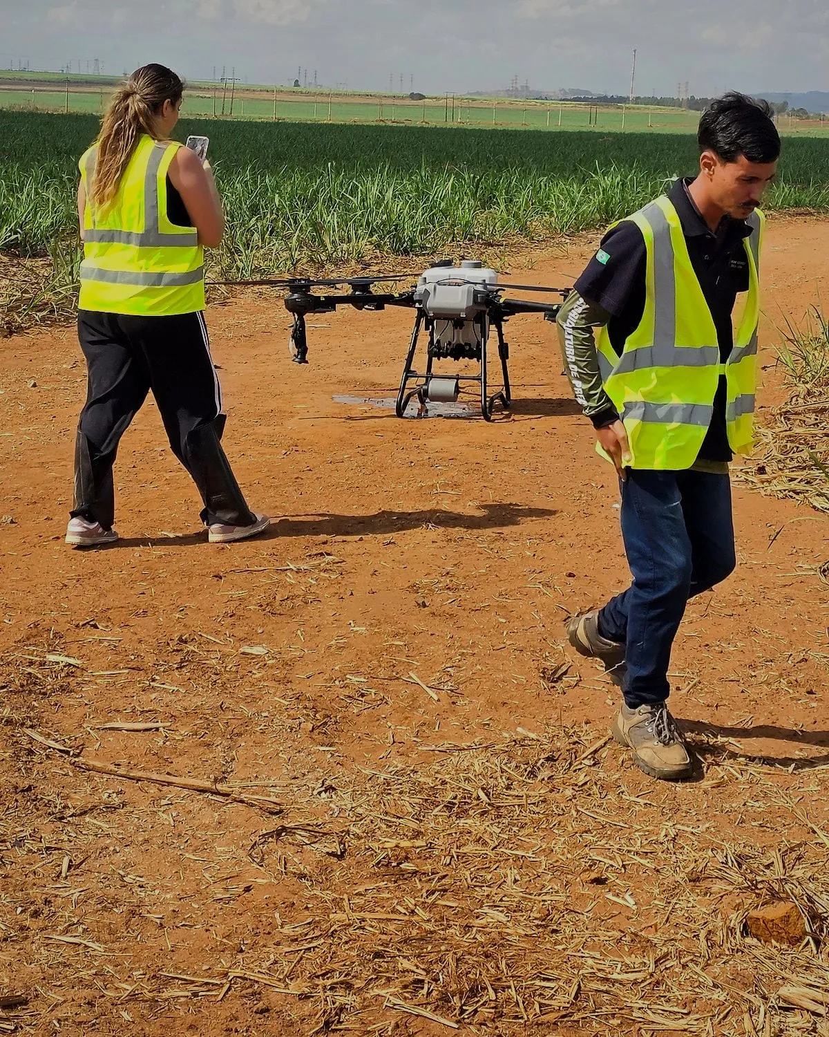Dois alunos de colete refletivo caminham ao lado de um drone DJI Agras pousado no campo