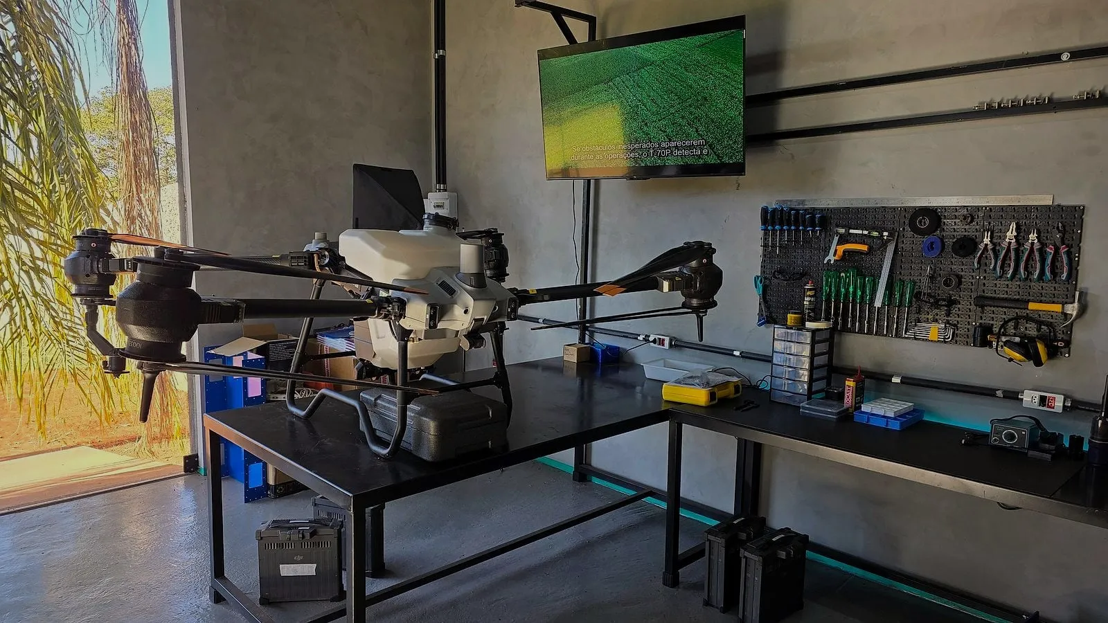 Drone DJI Agras sobre a bancada do laboratório da Drone Sense, com o painel de ferramentas e a TV ao fundo