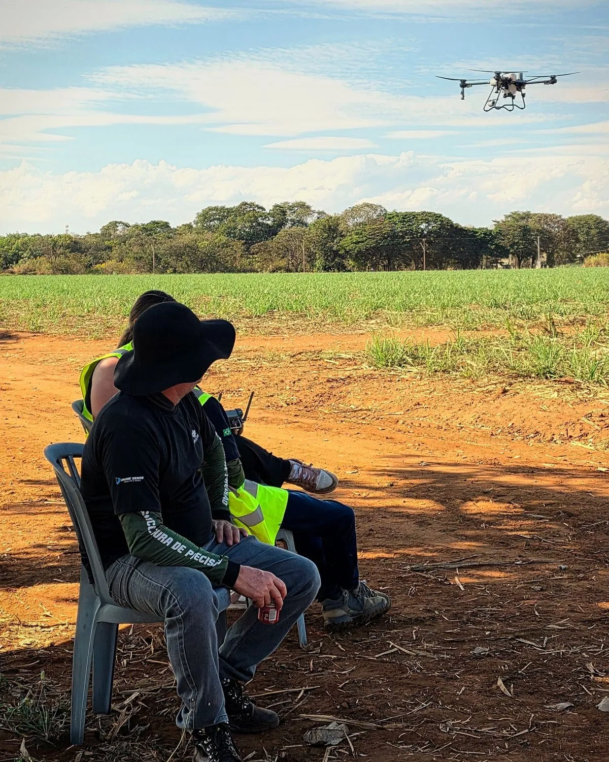 Instrutor de chapéu e aluno de colete sentados na sombra, de lado para a câmera, acompanhando um drone DJI Agras que voa sobre a lavoura