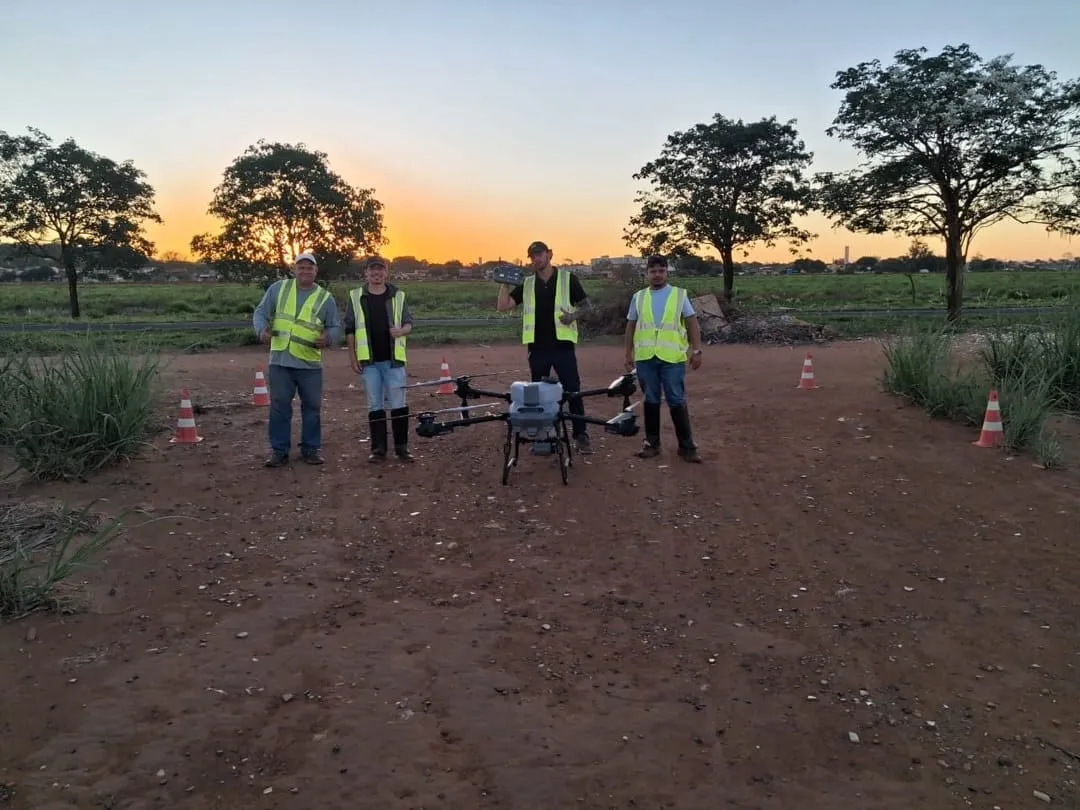 Quatro pessoas com colete refletivo em campo de terra, ao entardecer, ao lado de um drone DJI Agras cercado por cones